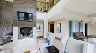 Located at 1701 Atria Cir in Raleigh, NC, this contemporary residence makes an immediate impression with its dramatic double-height living area, where soaring ceilings draw the eye upward to an open mezzanine level with sleek metal railing — a striking architectural focal point. The living space is anchored by a floor-to-ceiling stacked stone accent wall housing a wall-mounted television above a fireplace, lending a bold, textural contrast to the otherwise crisp, white interior. Light-toned flooring and neutral walls create a bright, airy atmosphere throughout, while curated contemporary artwork in cool blue and white tones adds a refined decorative touch. Plush light gray seating arranged around a low-profile coffee table encourages comfortable, casual living. Recessed lighting ensures consistent illumination across the open-plan layout. A deep-set nook within the stone wall offers a cozy, intimate alcove perfect for reading or quiet retreat. Hallways branch off toward additional rooms, providing a functional flow between spaces. The overall design aesthetic is modern and sophisticated, balancing clean architectural lines with warm textural elements to create a polished yet inviting living environment.
