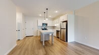 This single-floor residence features a clean, contemporary design highlighted by a cohesive neutral color palette and wood-look flooring throughout the main living areas. The open-concept kitchen serves as a focal point, showcasing white shaker-style cabinetry, light quartz countertops, and a subtle gray subway tile backsplash. A full suite of stainless steel appliances—including a refrigerator, electric range, microwave, and dishwasher—provides functional appeal, while a central island with a wood-paneled base and pendant lighting above adds both workspace and visual interest. The kitchen flows naturally into the living area, which features smooth hardwood-style flooring and a sliding glass door that opens to a private balcony with views of surrounding greenery. The bedroom is carpeted for comfort and receives natural light through a large window. Adjacent to the bedroom, the full bathroom includes a single-sink vanity with warm wood cabinetry, a tub-shower combination, and wood-look tile flooring. A dedicated closet area with built-in shelving provides practical storage. Throughout, recessed lighting and crisp white walls maintain a bright, well-finished ambiance.