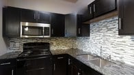 This single-floor space showcases a thoughtfully designed interior with a standout kitchen at its heart. The kitchen is finished with rich, dark espresso cabinetry that extends from floor to ceiling, offering generous storage through both upper and lower cabinets accented with sleek bar-pull hardware. The countertops feature dark granite with subtle veining, providing a durable and elegant work surface that pairs beautifully with the deep cabinetry tones. A stainless steel double-basin sink with a modern arc faucet is set beneath the countertop, while stainless steel appliances — including a range, over-the-range microwave, and oven — complete the contemporary aesthetic. The eye-catching mosaic tile backsplash runs the full length of the kitchen walls, showcasing a linear pattern in muted earth tones and soft greens that adds texture and visual interest to the space. Crown molding along the upper walls introduces a refined architectural detail, bridging the modern kitchen design with classic finishing touches. The overall ambiance is polished and cohesive, blending functionality with a sophisticated, contemporary style suited for everyday living and entertaining alike.