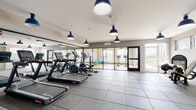 This fitness center is a thoughtfully designed, single-floor facility built to support a full range of workout routines. The space is finished with durable rubber-tile flooring throughout, providing both safety and comfort underfoot. Distinctive navy blue pendant lights hang from the white ceiling, lending the gym a modern, polished aesthetic while delivering ample overhead illumination.

The main workout area is lined with large wall mirrors that amplify the sense of space and allow users to monitor their form. A row of treadmills and elliptical cross-trainers occupies one section of the floor, each machine equipped with an individual display screen. Adjacent to the cardio equipment, a dedicated strength-training zone houses a cable machine, adjustable benches, and a well-stocked dumbbell rack offering a graduated range of weights.

Floor-to-ceiling glass partition walls divide the space into distinct zones, including what appears to be a flexible studio area appointed with exercise balls and additional equipment. This studio section benefits from direct access to natural light through large windows and a glass door that opens to outdoor views, creating an airy, motivating atmosphere.

The facility also features a separate entry with double exit doors, ensuring smooth traffic flow. Overall, the gym combines a clean, contemporary design with a comprehensive selection of equipment, making it a functional and inviting space for users of all fitness levels.