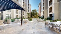 Located in the heart of Houston's prestigious River Oaks neighborhood, this outdoor amenity courtyard at 2900 W Dallas Street offers a thoughtfully designed retreat within a multi-story residential community. The space is anchored by a generously sized swimming pool with crystal-clear blue water, framed by a broad stone-tiled sun deck outfitted with contemporary chaise lounges and side tables — ideal for relaxing under the Texas sun. Mature trees provide natural shade across portions of the deck, creating a balance of open sunlight and sheltered comfort. Lush, meticulously maintained landscaping, including trimmed hedgerows, ornamental grasses, and tropical plantings, weaves throughout the courtyard, lending a verdant, resort-like atmosphere. A dedicated outdoor dining area features round tables with cushioned chairs beneath a sleek pergola structure, accompanied by a brick-built outdoor grilling station — a perfect setup for casual gatherings. The surrounding multi-story building façade, finished in warm stone and brick, provides an elegant architectural backdrop while enclosing the space to create an intimate, private ambiance. Natural light floods the courtyard throughout the day, enhancing the open, airy character of this well-appointed outdoor living space.