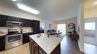 This single-floor residence showcases a thoughtfully designed open-concept layout that seamlessly integrates the kitchen, living area, and bedroom. The kitchen is outfitted with rich, dark espresso cabinetry accented by sleek hardware, a granite-topped island that provides ample prep and gathering space, and stainless steel appliances including a built-in microwave. Under-cabinet task lighting and a skylight-style fixture illuminate the workspace effectively.

The kitchen flows naturally into a spacious living area finished with warm wood-look flooring, which adds continuity throughout the space. The living room accommodates generous seating arrangements and features a large sliding glass door that invites natural light and provides access to an outdoor patio area. A bedroom is visible adjacent to the main living space, offering a private retreat with its own defined area.

The overall ambiance is modern and functional, with neutral wall tones complementing the contrasting dark cabinetry and light countertops. The open layout enhances the sense of space, making the unit feel airy and well-connected throughout its single level.