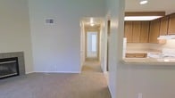 This single-floor residence offers a clean, functional layout designed for comfortable everyday living. The main living area features neutral-toned carpeting and smooth walls painted in a soft gray-green hue, creating a calm and understated ambiance throughout. A central hallway extends from the living area, connecting the various rooms of the home and providing a clear, organized flow between spaces. Flush-mount ceiling lighting illuminates the corridor, guiding the eye toward a bright room at the far end, suggesting ample natural light deeper within the unit. To one side of the living area, the kitchen is partially visible, showcasing warm wood-toned cabinetry, a white refrigerator, and a raised countertop bar that separates the kitchen from the main living space — a practical feature that promotes an open, connected feel while maintaining a defined cooking zone. Crisp white baseboards and door trim add a clean contrast against the soft wall tones, while a ceiling-mounted HVAC vent reflects thoughtful climate control integration. The overall design is practical and straightforward, offering a well-connected single-level floor plan suited for a variety of living arrangements.
