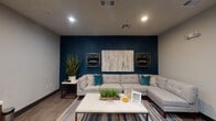 This single-floor space is designed as a sophisticated communal lounge area, thoughtfully divided into multiple distinct zones that together create a cohesive and welcoming environment. The layout features an open-plan design with wood-look tile flooring throughout, recessed ceiling lighting, and visible HVAC vents integrated cleanly into the ceiling.

A standout feature is the deep navy blue accent wall anchoring one of the primary lounge areas. This bold wall is adorned with a large abstract canvas painting flanked by two gold geometric circular wall shelves, lending the space an artistic, curated feel. A generously sized light grey L-shaped sectional sofa, accented with teal throw pillows, faces a white rectangular coffee table atop a multi-toned striped area rug, creating an inviting and cohesive seating arrangement.

The broader open lounge area incorporates a tufted white sofa with orange accent pillows, a glass-and-gold coffee table, and a dark upholstered armchair. A bold orange structural column adds a striking architectural pop of color. Glass-walled rooms are visible beyond, offering visual connectivity while maintaining separation. A wall-mounted flat-screen television with a wood media console anchors another seating zone. Tall decorative vases and lush indoor plants add warmth and organic texture throughout.