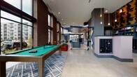 This expansive single-floor amenity space combines industrial and contemporary design elements to create a vibrant, multifunctional environment. Soaring double-height ceilings and floor-to-ceiling windows along multiple walls flood the interior with abundant natural light while offering open views of the surrounding outdoor landscape. Exposed brick columns serve as dramatic architectural anchors throughout, contrasting beautifully with the polished large-format tile flooring below.

The lounge area features plush emerald-green velvet sofas paired with mustard and rust-colored accent chairs arranged around metallic drum coffee tables, creating a welcoming social hub. Adjacent to the seating area, a full-size pool table positioned atop a geometric-patterned area rug provides a focal point for recreation.

Toward the interior, a fully equipped communal kitchen showcases slate-blue cabinetry, a subway tile backsplash, and stainless steel appliances, accented by a bold zigzag blue graphic column that adds artistic energy. A nearby mailroom area features built-in shelving, individual mailboxes, and a dramatic pop-art–inspired wallcovering depicting repeating golden faces against a dark background. Pendant lighting and track fixtures complete the eclectic, design-forward aesthetic throughout.