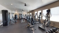 This single-story fitness center offers a comprehensive workout environment designed to accommodate a variety of exercise routines. The space is generously sized, providing ample room for members to move freely between stations without feeling crowded. The flooring is finished in a durable dark rubber material, ideal for high-traffic gym use and equipment stability.

Along one side of the room, a row of cardio machines — including treadmills, ellipticals, and stair climbers — lines the wall, several equipped with individual display screens for an enhanced workout experience. Strength-training equipment, including a multi-function cable machine and various weight benches with free weights, occupies the opposite side, creating a clear division between cardio and resistance training zones.

Natural light filters in through a series of white-trimmed windows fitted with blinds, brightening the interior alongside recessed ceiling lights. Multiple ceiling fans are evenly distributed throughout, ensuring consistent airflow and comfort during workouts. Neutral-toned walls contribute to a clean, focused atmosphere, while wall-mounted mirrors near the strength area support proper form and add to the sense of openness.