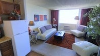 This single-floor space presents a thoughtfully designed interior that blends contemporary style with functional living. The living area serves as the focal point, featuring sleek white tufted sofas accented with houndstooth throw pillows, creating a chic and modern aesthetic. A bold rust-orange area rug with circular patterns anchors the seating arrangement, adding warmth and visual interest to the space. A dark coffee table with a glossy finish sits at the center, complementing the overall color palette. Warm ambient lighting is provided by elegant table and floor lamps, casting a cozy glow throughout the room. A large window dressed with rich burgundy curtains allows natural light to fill the space, enhancing its open and airy feel. A wall-mounted air conditioning unit ensures year-round comfort. The walls are kept neutral, allowing a vibrant triptych of modern artwork in shades of red, gray, and blue to serve as a striking decorative accent. The flooring transitions between hardwood and carpet, adding texture and dimension. Overall, the space exudes a stylish, well-curated ambiance that balances bold design choices with comfortable, everyday livability.