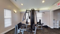 Located at 421 Iverson Way in Galloway, Ohio, this single-floor fitness facility offers a dedicated space for strength and resistance training. The workout room features vaulted ceilings that lend a sense of openness to the space, complemented by recessed lighting and wall-mounted sconces that ensure adequate illumination throughout. The room is finished with neutral-toned walls, white crown molding, and durable carpeted flooring designed to withstand the demands of an active fitness environment. Natural light filters in through multiple windows fitted with blinds, creating a bright and welcoming atmosphere. The centerpiece of the space is a robust multi-station cable and weight machine that supports a wide variety of upper and lower body exercises, including lat pulldowns, cable rows, and leg extensions. A set of free weights is also visible, providing additional options for traditional dumbbell training. The layout is thoughtfully arranged to allow users to move freely between stations. The overall ambiance is practical and purposeful, making this facility well-suited for residents or members seeking a convenient on-site option for maintaining an active fitness routine.