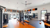 Located in Tomball, Texas, this fitness center is a thoughtfully designed exercise facility that delivers a comprehensive workout experience within a generously sized single-floor space. The interior is immediately distinguished by its bold orange and black color scheme, carried through the rubber flooring — a striking combination of orange and dark speckled zones — motivational wall murals, and branded signage promoting a "Mind, Balance, Body" philosophy.

The strength training area is well-appointed with a multi-functional cable machine, a Smith machine with adjustable bench, a dedicated dumbbell rack, and isolated strength machines, all arranged to allow ample circulation between equipment. Large floor-to-ceiling mirrors expand the visual space and allow users to monitor their form during exercises.

The cardio zone features recumbent bikes, rowing machines, and treadmills positioned near oversized windows that invite abundant natural light and provide outdoor views of the surrounding landscape — creating an open, energizing atmosphere. Ceiling fans throughout the space ensure comfort during intense workouts.

A functional training area rounds out the facility, stocked with stability balls, medicine balls, and kettlebells on a rolling rack. Recessed lighting, high white ceilings, and glass partition doors contribute to the airy, modern aesthetic, making this fitness center a fully equipped and visually motivating environment.
