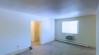 This single-floor residential space offers a straightforward and functional layout designed for comfortable everyday living. The main living area is finished with wall-to-wall carpet, providing a soft and practical underfoot surface throughout. Neutral-toned walls create a clean, bright atmosphere that allows for versatile interior styling.

Climate control is well-equipped, featuring both a wall-mounted air conditioning unit and a baseboard heating system, ensuring year-round comfort regardless of the season. A large window fitted with vertical blinds allows natural light to filter into the space while offering privacy and light control.

An interior doorway leads to an adjoining room, adding to the functional flow of the layout. The doorway opens into a warmly lit secondary space, suggesting additional utility such as a bedroom, bathroom, or storage area.

The overall ambiance of the space is understated and practical, with clean lines and a neutral palette that serves as an ideal canvas for personalization. The single-floor design ensures ease of movement throughout, making the space accessible and straightforward to navigate. This unit is well-suited for those seeking a no-frills, move-in-ready environment with essential amenities already in place.