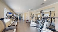 This single-story fitness center is designed to provide a thorough and functional workout environment. The space is laid out in an elongated, open floor plan that accommodates a wide variety of exercise equipment without feeling crowded. Durable dark concrete-style flooring runs throughout, offering a practical and low-maintenance surface suited for high-traffic use.

Cardio equipment is a central feature, with multiple treadmills, elliptical trainers, and stationary cycling bikes from the True Fitness brand arranged to allow ample space between machines. A multi-station cable and weight machine occupies the strength-training area, complementing an adjustable workout bench for free weight exercises. Large wall-mounted mirrors line portions of the room, enhancing the sense of openness and allowing users to monitor their form during workouts.

Recessed lighting and crown molding contribute a polished architectural finish, while multiple windows fitted with horizontal blinds admit natural light throughout the space. A glass-paned exterior door at one end opens to an outdoor area with a visible pool beyond, offering pleasant views and ventilation. An attached restroom provides added convenience. The overall ambiance is clean, bright, and purposefully organized for effective fitness use.