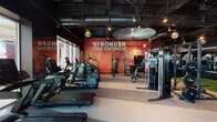 This single-floor fitness center delivers a comprehensive workout environment designed to inspire and perform. The space is anchored by a striking red mosaic accent wall emblazoned with the motivational phrase "Stronger Than Yesterday," setting an energetic tone throughout. The dark ceiling with exposed ductwork and industrial-style track lighting creates a sleek, modern aesthetic that contrasts effectively with the bold wall graphics.

The cardio zone features a lineup of treadmills, ellipticals, and stair climbers, with a dedicated section positioned directly in front of expansive floor-to-ceiling windows that look out onto a sunny outdoor terrace complete with palm trees and lounge seating, allowing natural light to flood the space during workouts.

The strength training area includes a full dumbbell rack with mirrors, functional training rigs equipped with TRX suspension straps, cable machines, and a multi-use power rack situated on a turf surface ideal for dynamic movements. Additional cable and resistance machines are distributed throughout the floor.

Geometric pendant lights add a contemporary design touch, while ceiling fans contribute to air circulation. The flooring transitions between rubber, carpet tile, and artificial turf zones, delineating distinct training areas. Large glass partitions offer views into adjacent amenity spaces, reinforcing the open, connected feel of this well-appointed fitness facility.