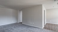 This single-floor residence on Pinewood Drive in Yorktown, Virginia, offers a thoughtfully laid-out interior finished in a neutral, light palette throughout. The open living area is generously sized, featuring plush gray carpeting and a large sliding glass door dressed with vertical blinds that allows natural light to pour in while providing access to an outdoor view. A dedicated entryway with wood-look flooring transitions seamlessly into the main living space.

The kitchen is equipped with black stainless steel appliances, including a gas range, over-the-range microwave, and a full-size refrigerator. Light granite-style countertops contrast the dark cabinetry, and wood-look flooring continues underfoot. The kitchen connects to an adjacent dining or utility area via an open passageway, adding to the functional flow.

The bedrooms are carpeted and filled with natural light from well-positioned windows. The primary bedroom features dual sets of bifold closet doors offering generous built-in storage with hanging rods and shelving. Additional bedrooms are equally spacious and versatile. The bathroom includes a white vanity with under-cabinet storage, a white countertop, and oil-rubbed bronze fixtures, presenting a clean, updated aesthetic.