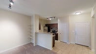 This single-floor space in Addison offers an open-concept layout that connects the living area to the kitchen, creating a cohesive and functional environment. The kitchen is outfitted with dark espresso cabinetry, a granite-topped breakfast bar, and stainless steel appliances including a refrigerator, microwave, and electric range. Tile flooring runs throughout the kitchen and entry areas, while plush carpeting covers the living space, providing a comfortable contrast underfoot. A set of built-in open shelves lines one wall adjacent to the kitchen, offering convenient display or storage options. Track lighting in the kitchen ensures the cooking area is well-lit, complementing the neutral, light-toned walls that give the space a clean and airy feel. A standard entry door is visible toward the rear of the kitchen area, and the overall layout makes efficient use of the available space. The open design between the kitchen and living area promotes an easy flow, making the space well-suited for everyday living and casual entertaining.