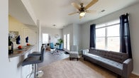 This single-floor space offers a well-designed, open-concept layout that seamlessly blends the living and dining areas into one cohesive and functional environment. The living area is anchored by soft carpeting, a tufted gray sofa, and a cushioned accent chair, all arranged around a central area rug that adds warmth to the space. A ceiling fan overhead provides both comfort and a finishing architectural touch. Large windows dressed with navy curtains flood the room with natural light and offer open views of the surrounding landscape, creating an airy and welcoming atmosphere.

Adjacent to the living area, the dining space features a round glass-top table paired with contemporary blue upholstered chairs, adding a pop of color that complements the neutral gray tones of the walls and flooring. A set of white French doors opens to the exterior, further enhancing the flow of natural light and providing convenient outdoor access.

The hard-surface flooring in the dining and entry areas transitions smoothly into the carpeted living zone, delineating spaces while maintaining visual continuity. A wall-mounted flat-screen television and sleek media console complete the living area. The overall ambiance is clean, modern, and inviting, with a neutral color palette that lends itself well to a variety of interior styles.