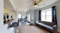 This single-floor space offers a well-designed, open-concept layout that seamlessly blends the living and dining areas into one cohesive and functional environment. The living area is anchored by soft carpeting, a tufted gray sofa, and a cushioned accent chair, all arranged around a central area rug that adds warmth to the space. A ceiling fan overhead provides both comfort and a finishing architectural touch. Large windows dressed with navy curtains flood the room with natural light and offer open views of the surrounding landscape, creating an airy and welcoming atmosphere.

Adjacent to the living area, the dining space features a round glass-top table paired with contemporary blue upholstered chairs, adding a pop of color that complements the neutral gray tones of the walls and flooring. A set of white French doors opens to the exterior, further enhancing the flow of natural light and providing convenient outdoor access.

The hard-surface flooring in the dining and entry areas transitions smoothly into the carpeted living zone, delineating spaces while maintaining visual continuity. A wall-mounted flat-screen television and sleek media console complete the living area. The overall ambiance is clean, modern, and inviting, with a neutral color palette that lends itself well to a variety of interior styles.