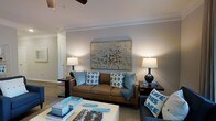 Located on Maybank Highway in Johns Island, South Carolina, this single-story space presents a well-appointed interior with a cohesive and inviting design aesthetic. The living area serves as the central gathering space, showcasing neutral gray walls that provide a versatile backdrop for the warm, layered furnishings throughout. Crown molding lines the ceiling, adding a refined architectural touch, while a ceiling fan contributes both style and function overhead. The room is anchored by a rich brown leather sofa adorned with blue and white patterned accent pillows, complementing the cool blue tones woven throughout the space. A large, framed abstract artwork commands attention on the main wall, flanked by matching table lamps that cast a warm, ambient glow. A patterned area rug grounds the seating arrangement, and a coffee table styled with decorative trays and accents adds polish to the overall composition. The flooring transitions smoothly toward an adjacent hallway, hinting at additional spaces beyond. The overall ambiance is warm, curated, and welcoming, blending comfort with tasteful design elements suited to the relaxed character of the Johns Island locale.