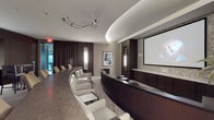 This single-floor space functions as a refined screening or presentation room, thoughtfully designed with both form and function in mind. The room is anchored by a large projection screen mounted against a textured stone-tile accent wall, paired with a ceiling-mounted projector that ensures a high-quality viewing experience. A curved, dark wood AV console spans the front of the room, providing a sleek and organized hub for audio-visual controls and equipment.

Seating is arranged in a gently curving configuration along one side, with plush white leather chairs offering comfort for extended use. The layout encourages an unobstructed sightline to the screen from every position. On the opposite side, a long dark conference-style table extends along the wall, suggesting the room can also serve as a hybrid boardroom or multi-purpose meeting space.

The ceiling features a graceful curved cove detail with integrated ambient lighting, contributing to the room's polished aesthetic. Warm-toned wall sconces provide supplemental lighting, while dark drapery along the far wall allows for full light control. Framed artwork adds a subtle decorative touch, balancing the room's professional atmosphere with a welcoming, contemporary ambiance.