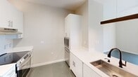 This thoughtfully designed one-bedroom, one-bathroom unit offers a modern and functional living experience across a single floor. The kitchen serves as a central highlight, featuring white cabinetry, light quartz countertops with an integrated stainless steel sink, stainless steel appliances including a refrigerator and range, a built-in microwave tower, and a sleek overhead linear pendant light that adds a contemporary industrial touch. The open counter layout provides practical workspace and a natural transition into the living area. The bedroom is a bright, comfortable space bathed in natural light from a large window, with neutral warm-toned walls, polished concrete-style flooring, and a generously sized closet fitted with wire shelving and double panel doors. The bathroom is finished to a high standard, showcasing a marble-look quartz vanity countertop with an undermount sink, matte black fixtures, a backlit LED vanity mirror, and a walk-in shower visible in the reflection. The unit also includes in-unit washer and dryer hookups, adding everyday convenience. Consistent polished concrete flooring runs throughout, tying the space together with a clean, modern aesthetic suited for contemporary living.