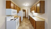 This one-bedroom, one-bathroom unit is thoughtfully laid out across a single floor, making efficient use of its cozy footprint. The galley-style kitchen is a standout feature, lined on both sides with warm oak cabinetry that extends to the ceiling for ample storage. It is equipped with a full suite of white appliances, including a coil-burner electric range, built-in microwave, refrigerator, and dishwasher, complemented by light-toned laminate countertops and a double-basin sink. Tile-pattern flooring runs throughout the kitchen, adding a practical, easy-to-clean surface. A ceiling fan with a light fixture provides airflow and illumination in the kitchen area. The bedroom offers comfortable carpeting underfoot, a ceiling fan with lighting, a window dressed with vertical blinds for natural light control, a baseboard heater, and a spacious reach-in closet with double doors and a full-width hanging rod. The bathroom is clean and functional, featuring a full tub and shower combo unit alongside standard fixtures and a vanity counter. A dedicated laundry room and connecting hallways add to the unit's practicality. Textured ceilings are consistent throughout, contributing to the space's classic residential character.
