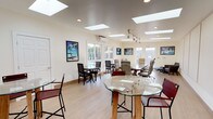 This single-floor space is thoughtfully designed as a versatile professional environment, combining open-plan flexibility with distinct functional zones. Multiple skylights are distributed across the ceiling, flooding the interior with consistent natural light and complementing the warm, light-toned hardwood flooring that runs throughout. Recessed lighting and adjustable track fixtures supplement the natural illumination, ensuring the space is well-lit at all hours.

A central conference area anchors the layout, featuring a substantial wood-and-glass table surrounded by ergonomic executive chairs. This area is ideal for formal meetings or collaborative work sessions. Acoustic wall panels line one section of the room, enhancing sound quality and adding a refined, professional finish. A wall-mounted mini-split system provides climate control.

Multiple informal seating areas are distributed around the perimeter, each featuring glass-top tables with chairs in varying styles, offering flexibility for breakout conversations or independent work. Large windows and glass-panel doors along the walls frame views of an outdoor area with a pool and surrounding greenery, bringing the outdoors in. Framed photographic artwork decorates the cream-toned walls, contributing a polished yet relaxed atmosphere throughout the space.