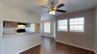 This single-story space showcases an open-concept layout that seamlessly connects the living, dining, and kitchen areas. The flooring throughout the main living space consists of rich, dark hardwood-style planks that contrast cleanly against the neutral gray walls and bright white baseboards, lending the space a polished, contemporary feel.

The living and dining area is illuminated by natural light streaming through multiple windows fitted with white horizontal blinds, creating a bright and welcoming atmosphere. A ceiling fan with an integrated light fixture is centrally positioned overhead, providing both airflow and additional lighting. The textured popcorn ceiling adds a classic residential touch.

The kitchen is accessible via a pass-through counter that opens directly into the main living area, promoting a connected and sociable layout. Gray cabinetry lines the kitchen walls, complemented by light-toned countertops and a stainless steel sink. A range with an overhead microwave or hood is also visible, completing the functional kitchen setup.

Overall, the space offers a clean, functional design with a neutral color palette, making it highly adaptable to various interior styles.