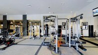 This modern fitness center offers a comprehensive workout environment designed to accommodate a wide range of training styles and fitness goals. The space is thoughtfully laid out on a single floor, with dedicated zones for strength training, functional fitness, and conditioning. At the center of the room, a professional Life Fitness multi-function cable and strength machine serves as the focal point, complemented by additional weight training equipment, free weights, and a heavy punching bag for combat-style training. Battle ropes and resistance accessories are readily accessible for functional and high-intensity workouts. A medicine ball rack lines one wall, offering a variety of weighted options for dynamic exercises. The interior design strikes a clean, contemporary aesthetic, with recessed lighting in a smooth white ceiling that keeps the space bright and energizing. Large windows or glass partitions allow natural light to filter in, enhancing the open, airy feel of the room. Dark stone accent columns add an architectural contrast to the otherwise light and neutral palette. Durable, patterned carpeting with defined grid lines helps visually organize the workout areas. The overall ambiance is polished and professional, reflecting a well-maintained facility suited for serious fitness enthusiasts.