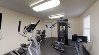 Located at 815 Cole Dr in Johnstown, Ohio, this single-floor fitness facility offers a dedicated space designed to support a well-rounded exercise routine. The workout room is outfitted with a selection of cardio equipment, including an elliptical trainer and a recumbent exercise bike, providing low-impact options for cardiovascular conditioning. A multi-station cable and weight machine anchors the strength training area, offering a variety of resistance exercises within a compact footprint. The flooring throughout the exercise area is finished in durable dark rubber matting, a practical choice that provides cushioning and traction during workouts. The room is well-lit by flush-mounted overhead panel lights set against a textured ceiling, creating a bright and functional environment. The neutral, light-colored walls contribute to an open and clean atmosphere. A private door provides access to the space, ensuring a self-contained area suited for focused fitness activity. Overall, the facility is efficiently arranged to maximize usable floor space while accommodating both cardiovascular and strength training needs in a straightforward, no-frills setting.