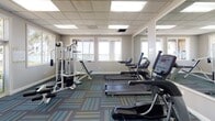 Located at 555 Lakeview Dr in Coral Springs, Florida, this well-appointed fitness center offers a comprehensive single-floor layout designed to support a full range of workout routines. The space is bright and welcoming, with rows of large windows along multiple walls that flood the interior with natural light and provide pleasant views of the surrounding outdoor amenity areas, including glimpses of a pool and lush landscaping.

The main workout area is equipped with a selection of cardio machines, including treadmills and elliptical trainers from True Fitness, as well as a multi-station strength training machine and a stationary bike. A wall-mounted flat-screen television enhances the cardio experience. Full-length mirrors are positioned along one wall, adding depth to the space and supporting proper form during exercise.

A separate, open flex area — lined with a bank of windows overlooking the outdoor grounds — provides ample room for stretching, yoga, or floor-based workouts. The facility features stylish patterned carpet tiles in teal, blue, and green tones throughout, complemented by neutral gray walls with green accent walls that create an energetic yet focused atmosphere. A suspended acoustic tile ceiling with recessed panel lighting ensures consistent, comfortable illumination. Glass-paneled double doors provide easy access between areas and to adjacent amenity spaces.