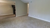 This single-floor space presents a clean, functional layout with a comfortable living area as its centerpiece. The room features plush, neutral-toned carpeting that adds warmth underfoot, while the walls are finished in a soft greige tone complemented by crisp white baseboards throughout. A notable architectural detail is the vaulted ceiling, which lends a sense of openness and vertical dimension to the living space.

An open passageway connects the carpeted living area to the kitchen, where the flooring transitions to hardwood-style planks. The kitchen is fitted with dark espresso cabinetry that provides a striking contrast against the lighter wall tones, offering ample storage. The ceiling-mounted HVAC vents are integrated throughout, ensuring efficient climate control across the space.

The overall ambiance is understated and move-in ready, with a neutral color palette that offers flexibility for personalization. The open sightlines between rooms enhance the sense of flow and connectivity, making the layout feel cohesive and practical for everyday living.
