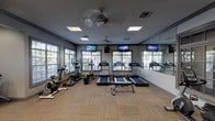 This fitness center is organized across a single floor and divided into distinct zones designed to support a full range of workout routines. The cardio area is generously appointed with multiple treadmills, elliptical trainers, and a stationary bike, each equipped with individual digital displays. Multiple flat-screen TVs are mounted on the walls, and large windows fitted with blinds line the perimeter, filling the space with natural light and offering views of surrounding greenery. A full-wall mirror in the cardio zone further enhances the sense of openness. The flooring throughout features a warm parquet-style wood pattern, adding a polished, welcoming feel underfoot. Ceiling fans provide additional climate comfort. An adjoining strength training area offers a multi-station cable machine, a weight bench with a leg press attachment, and a rack of dumbbells suited for a variety of resistance exercises. A dedicated functional training nook, distinguished by its deep teal accent wall, houses stability balls in assorted sizes, resistance bands, medicine balls, and other versatile accessories. The overall design balances practicality with a clean, modern aesthetic, creating an inviting environment conducive to consistent training across all fitness levels.