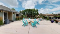 This single-level outdoor amenity space serves as a central gathering area within a residential community. The focal point of the courtyard is a round, stone fire pit encircled by six turquoise Adirondack chairs accented with colorful patterned cushions, creating a lively and inviting atmosphere. The spacious concrete patio extends generously in multiple directions, providing ample room for residents to move freely between seating zones.

To one side, a sectional outdoor sofa in neutral tones offers an additional lounge area, while a separate dining-style seating arrangement provides a more structured option for small groups. The courtyard is framed by carefully maintained landscaping, including ornamental grasses, shrubs, and potted plants that add texture and greenery along the borders. A decorative gravel border with curved edging runs along the building's exterior, adding a polished, finished look.

The space is accessed through a set of modern double glass doors adorned with botanical artwork, offering a clean and welcoming entry point. The exterior of the building features horizontal lap siding in a crisp off-white tone. Views of surrounding multi-story residential buildings and mature trees are visible beyond an iron perimeter fence, contributing to a sense of community while maintaining an open, airy feel under wide open skies.