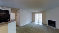 This single-floor space features an open-concept layout that connects the living area to the kitchen. The living room is finished with wall-to-wall carpeting and neutral-toned walls, creating a clean and simple aesthetic throughout. A textured ceiling adds subtle visual interest overhead, while a ceiling fan with integrated lighting provides both airflow and ambient illumination.

A fireplace anchors one side of the living room, offering a functional focal point. On the opposite end, a sliding glass door fitted with vertical blinds allows natural light to filter into the space, brightening the room considerably. This door likely provides access to an outdoor area.

The kitchen is partially visible and features a countertop overhang that separates it from the living space, along with a dark-toned countertop surface and a sink. Doorways visible from the living area lead to additional rooms, suggesting a practical and navigable floor plan. A ceiling-mounted light fixture near the entryway provides additional lighting in the transitional areas. Overall, the space offers a straightforward, functional design with neutral finishes throughout.