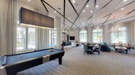 This single-story clubhouse is designed to serve as a sophisticated community gathering space, combining elegant aesthetics with versatile amenities. The main social hall is expansive and open, anchored by soaring ceilings accented with a distinctive geometric dark-beam grid pattern and recessed lighting that illuminates the entire area. Large windows dressed with sheer floor-to-ceiling drapes line the walls, allowing abundant natural light and views of the surrounding greenery to filter throughout.

The lounge area is furnished with an eclectic mix of patterned armchairs, side tables, and console tables, creating comfortable conversation zones. A wall-mounted flat-screen television and a decorative chandelier add both function and visual interest. Adjacent to the lounge, a full billiards table serves as a central recreational feature, accompanied by a large pendant light overhead.

A wet bar and catering kitchen are equipped with granite countertops, a double sink, beverage refrigerators, a mounted television, and decorative wall-mounted sculptural pieces, making it well-suited for hosting events. A smaller dining nook with high-top seating and its own wall-mounted screen provides a casual dining or meeting area. Throughout, striped carpeting, neutral tones, and carefully curated décor contribute to a refined yet welcoming ambiance.