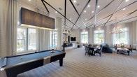 This single-story clubhouse is designed to serve as a sophisticated community gathering space, combining elegant aesthetics with versatile amenities. The main social hall is expansive and open, anchored by soaring ceilings accented with a distinctive geometric dark-beam grid pattern and recessed lighting that illuminates the entire area. Large windows dressed with sheer floor-to-ceiling drapes line the walls, allowing abundant natural light and views of the surrounding greenery to filter throughout.

The lounge area is furnished with an eclectic mix of patterned armchairs, side tables, and console tables, creating comfortable conversation zones. A wall-mounted flat-screen television and a decorative chandelier add both function and visual interest. Adjacent to the lounge, a full billiards table serves as a central recreational feature, accompanied by a large pendant light overhead.

A wet bar and catering kitchen are equipped with granite countertops, a double sink, beverage refrigerators, a mounted television, and decorative wall-mounted sculptural pieces, making it well-suited for hosting events. A smaller dining nook with high-top seating and its own wall-mounted screen provides a casual dining or meeting area. Throughout, striped carpeting, neutral tones, and carefully curated décor contribute to a refined yet welcoming ambiance.