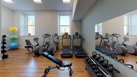 This fitness center is a thoughtfully designed, single-floor space that offers a comprehensive range of workout equipment to support diverse training goals. The room features warm hardwood flooring throughout, clean white and sage-green walls, and a combination of large windows and recessed lighting that create a bright, energizing atmosphere with views of the surrounding area.

The cardio zone is well-stocked with multiple treadmills, elliptical trainers, and a stationary bike, arranged to make efficient use of the space while allowing comfortable movement between machines. Large mirrors line one wall, amplifying the sense of space and allowing users to monitor their form during workouts.

The strength training area is equally well-appointed, featuring a multi-functional cable machine, a dedicated weight bench, and a comprehensive dumbbell rack spanning a wide range of weights. Kettlebells and medicine balls round out the free-weight offerings, providing ample options for varied training routines.

The layout is open and functional, with high ceilings in the main strength area adding a sense of spaciousness. Storage closets are neatly integrated into the design, maintaining a clean and organized environment. The overall ambiance is professional and welcoming, making this fitness center a well-rounded facility suitable for both cardio-focused and strength-based workouts.