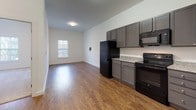 This 1-bedroom, 1-bathroom unit is thoughtfully designed to maximize functionality across a generous single-floor layout. The open-concept living and kitchen area is anchored by warm wood-look flooring that runs throughout, creating a cohesive and inviting atmosphere. The kitchen features grey shaker-style cabinetry, granite-look countertops, and a full suite of black appliances, including a refrigerator, range, built-in microwave, and dishwasher. Ample upper and lower cabinet storage ensures plenty of room for organization, while the L-shaped layout provides efficient use of space. The living area flows naturally from the kitchen, offering generous room for furnishings and everyday living. The bedroom is carpeted for comfort and features a large window that allows natural light to fill the space, contributing to a bright and airy feel. The bathroom is clean and modern, appointed with tile flooring, a dark-toned vanity with granite-look countertop, an undermount sink, and a framed mirror with overhead lighting. White walls throughout the unit create a neutral, move-in-ready canvas that complements the contemporary finishes. The layout is practical and well-proportioned, making it an efficient and comfortable living space.