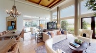 This single-story amenity space is designed to impress, combining architectural sophistication with a welcoming, resort-like atmosphere. The main lounge area commands attention with soaring ceilings accented by exposed wooden beams and an expansive wall of floor-to-ceiling windows and glass panels that frame sweeping views of a manicured outdoor landscape, including a pool, mature trees, and lush greenery. Natural light floods the interior, creating a bright and airy ambiance throughout.

The open layout features a spacious seating area furnished with neutral-toned sofas, accent chairs, and layered area rugs, anchored by a large flat-screen television. A rotunda-style entry area with a decorative column, coved ceiling, and an ornate chandelier adds elegance to the transition between zones. Built-in display niches and a circular wall mirror contribute refined decorative touches.

A dedicated media or theater room is accessed through a standout stone-surround archway fitted with solid wood double doors, offering an intimate retreat within the space. A well-equipped refreshment station, featuring bold crimson accent walls, a commercial beverage machine, stone countertops, and under-counter storage, provides convenient service functionality. The combination of natural materials, curated furnishings, and abundant outdoor views gives this space a polished, community-focused character.