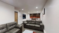 This versatile basement area seamlessly combines a comfortable living space with a functional kitchenette. The room is illuminated by recessed lighting, creating a warm ambiance despite the lack of natural light. The lounge area is furnished with plush leather seating, including a three-seater sofa and an armchair, both in a rich gray tone. These pieces are positioned on a patterned area rug, adding texture to the space.

The kitchenette, visible at the far end of the room, features dark wood cabinetry contrasted against a vibrant orange backsplash. This color pop adds energy to the otherwise neutral palette. A large flat-screen TV mounted on the white textured wall serves as a focal point, ideal for entertainment.

The room's design maximizes space efficiency, with a compact layout that doesn't sacrifice comfort. The white walls and ceiling help to brighten the space, while the wood-look flooring adds warmth. A wooden door, likely leading to other areas of the home, is visible, suggesting this basement space is well-integrated into the house's overall layout.