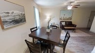 This single-story space presents an open-concept layout that seamlessly connects the dining and living areas, creating a cohesive and functional environment. The dining area is anchored by a dark-stained wood table paired with matching high-back chairs, offering comfortable seating for four. A tall window dressed with sheer curtains allows natural light to filter softly into the space, illuminating the neutral-toned walls and wood-look flooring that run throughout.

The adjacent living area is furnished with a dark leather sofa, an accent chair, and a patterned area rug that helps define the seating zone. A ceiling fan overhead provides both practical airflow and a subtle design element. Decorative wall art featuring landscape imagery adds visual interest and a touch of character to the space, while a floor lamp provides warm supplemental lighting.

The overall ambiance is casual yet well-appointed, with a cohesive color palette of warm neutrals and dark wood tones that tie the two areas together harmoniously. The open flow between the dining and living zones makes the space feel inviting and well-suited for both everyday living and relaxed entertaining.