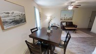 This single-story space presents an open-concept layout that seamlessly connects the dining and living areas, creating a cohesive and functional environment. The dining area is anchored by a dark-stained wood table paired with matching high-back chairs, offering comfortable seating for four. A tall window dressed with sheer curtains allows natural light to filter softly into the space, illuminating the neutral-toned walls and wood-look flooring that run throughout.

The adjacent living area is furnished with a dark leather sofa, an accent chair, and a patterned area rug that helps define the seating zone. A ceiling fan overhead provides both practical airflow and a subtle design element. Decorative wall art featuring landscape imagery adds visual interest and a touch of character to the space, while a floor lamp provides warm supplemental lighting.

The overall ambiance is casual yet well-appointed, with a cohesive color palette of warm neutrals and dark wood tones that tie the two areas together harmoniously. The open flow between the dining and living zones makes the space feel inviting and well-suited for both everyday living and relaxed entertaining.