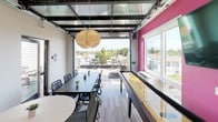 This dynamic rooftop amenity space is thoughtfully designed to blend indoor comfort with outdoor living. At its heart is a main gathering room featuring a striking glass garage door that opens fully to an expansive outdoor terrace, seamlessly merging the two areas. The interior is characterized by bold design choices, including a vibrant magenta accent wall, light wood-style flooring, and a sculptural globe pendant light that anchors the space visually.

A full-length shuffleboard table runs along one wall, accompanied by a wall-mounted flat-screen television, making it an ideal setting for recreation and entertainment. A kitchenette area provides practical amenity support, featuring dark cabinetry with a light countertop and a striking hexagonal patterned tile backsplash in muted tones. High-top bistro seating and a dining table offer flexible arrangements for both casual and group gatherings.

The outdoor terrace extends the living space considerably, offering open-air seating with high-top tables, a gas grill, and a glass railing system that preserves sweeping views of the surrounding neighborhood and city skyline beyond. A glass roof canopy over the interior transition area allows natural light to pour in, further brightening the space. Together, the indoor and outdoor components create a well-appointed, multi-functional environment suited for social events and everyday enjoyment.