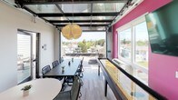 This dynamic rooftop amenity space is thoughtfully designed to blend indoor comfort with outdoor living. At its heart is a main gathering room featuring a striking glass garage door that opens fully to an expansive outdoor terrace, seamlessly merging the two areas. The interior is characterized by bold design choices, including a vibrant magenta accent wall, light wood-style flooring, and a sculptural globe pendant light that anchors the space visually.

A full-length shuffleboard table runs along one wall, accompanied by a wall-mounted flat-screen television, making it an ideal setting for recreation and entertainment. A kitchenette area provides practical amenity support, featuring dark cabinetry with a light countertop and a striking hexagonal patterned tile backsplash in muted tones. High-top bistro seating and a dining table offer flexible arrangements for both casual and group gatherings.

The outdoor terrace extends the living space considerably, offering open-air seating with high-top tables, a gas grill, and a glass railing system that preserves sweeping views of the surrounding neighborhood and city skyline beyond. A glass roof canopy over the interior transition area allows natural light to pour in, further brightening the space. Together, the indoor and outdoor components create a well-appointed, multi-functional environment suited for social events and everyday enjoyment.