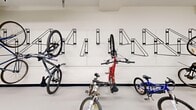 Located at 255 Carrington Blvd NW in Calgary, Alberta, this dedicated bike storage cage offers a practical and well-organized space for residents to securely store their bicycles. The single-level room is finished with clean white walls, a smooth sealed floor, and a dark base trim that gives the space a tidy, functional aesthetic.

The room is equipped with numerous wall-mounted bike racks installed along all perimeter walls, designed to hold bicycles both vertically and at an angle, maximizing storage capacity while keeping the floor area clear and accessible. The layout accommodates a variety of bicycle sizes, from full-sized mountain and road bikes to smaller children's bikes and tricycles.

Overhead, the space is illuminated by bright fluorescent strip lighting, ensuring excellent visibility throughout. Exposed utility ducting runs along the ceiling, reflecting the utilitarian character of the space. Two secure access doors — each fitted with electronic locks and emergency exit hardware — provide convenient entry and exit points from different sides of the room.

Safety features include a fire alarm system and security camera, ensuring the space is both monitored and protected. Overall, this bike cage provides a clean, secure, and efficiently designed storage solution for residents at this Calgary address.