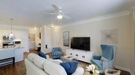 This single-floor residence offers a thoughtfully designed living space that blends comfort and style. The living room serves as the heart of the home, featuring crown molding along the ceiling, a ceiling fan with integrated lighting, and warm hardwood flooring that anchors the space with a timeless appeal. Decorative columns add an architectural touch, subtly defining the open layout while maintaining a sense of flow throughout the interior. The room is generously sized, accommodating a plush seating arrangement centered around a large flat-screen television mounted on a sleek white media console. Blue accent chairs and coordinating throw pillows lend the space a cohesive coastal-inspired palette that feels both inviting and polished. Natural light filters into the room, brightening the neutral wall tones and enhancing the overall sense of openness. Visible from the living area, an adjacent laundry space with stacked or side-by-side units adds a practical convenience. The front entryway is clearly defined with a paneled door and coat storage nearby. Overall, this residence presents a clean, well-maintained interior that balances functional living with refined decorative details throughout.