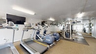 Located at 4877 Columbia Rd in North Olmsted, Ohio, this single-floor fitness center offers a well-organized workout facility across a compact but efficiently utilized footprint. The space is divided into distinct functional zones, each designed to support a comprehensive exercise routine.

The main workout area is equipped with multiple treadmills, an elliptical trainer, and a stair climber, all arranged to maximize floor space. A large wall mirror with motivational "No Pain No Gain" signage spans one side of the room, amplifying the sense of space and adding an energizing atmosphere. Protective rubber floor mats cover the strength training zone, which features a multi-cable weight machine suitable for a variety of resistance exercises. A dumbbell rack provides additional free-weight options.

A large window fitted with vertical blinds allows natural light to filter into the cardio area, creating a bright and welcoming environment. Overhead fluorescent lighting ensures consistent illumination throughout. The floors are finished with neutral carpeting in the transitional and stretching areas, providing comfort underfoot.

A dedicated stretching and mat room offers yoga mats on a storage rack, along with motivational wall decals. An entryway area includes a coat closet with hanging rods and a cushioned bench, providing a convenient transition space. The textured ceilings and clean gray walls maintain a functional, no-frills aesthetic throughout.