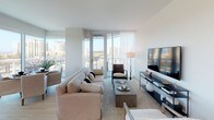 Unit #702 at 8800 Lombard in San Diego is a thoughtfully designed model residence that exemplifies contemporary urban living. The open-concept living area is anchored by expansive floor-to-ceiling windows and sliding glass doors that frame sweeping views of the San Diego skyline and lush treetops, flooding the space with abundant natural light. Light wood flooring flows throughout, lending warmth and continuity to the clean, neutral palette.

The kitchen is outfitted with stainless steel appliances, including a gas range and built-in microwave, complemented by crisp white cabinetry, quartz countertops, and a marble-style backsplash. A large picture window above the sink frames a serene greenery view, making the space as functional as it is visually appealing.

The bedrooms are generously proportioned, each offering large windows or sliding glass doors with city and treetop views. The primary suite connects seamlessly to a sleek en-suite bathroom featuring illuminated vanity mirrors, a white undermount sink, and an adjacent walk-in closet with built-in shelving and hanging storage. A secondary bathroom includes a soaking tub with tiled surround and a separate vanity area. Throughout the unit, the design emphasizes clean lines, soft tones, and a refined, modern aesthetic.