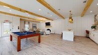 Located in Savannah, Georgia, this single-floor amenity facility seamlessly blends social and wellness spaces into one cohesive destination. The lounge and game room area immediately impresses with its exposed natural wood ceiling beams, light wood-look flooring, and crisp white board-and-batten accent walls. A central fireplace with a warm wood mantel and wall-mounted TV serves as the focal point of the seating area, complemented by a modern chandelier overhead. A full-size pool table anchors the game area, while a nearby wet bar features vibrant tropical tilework, open shelving, a beverage refrigerator, and a stylish island with bar seating — all accented by woven pendant lighting and arched windows that bring in natural views of the surrounding greenery. The fitness center is expansive and thoroughly equipped, housing treadmills, an elliptical, cable machines, free weights, adjustable benches, medicine balls, battle ropes, a heavy bag, and a multi-functional training rig. Floor-to-ceiling mirrors, ceiling fans, and arched windows keep the space bright and well-ventilated. A dedicated hydration station with dual water fountains and a bottle-fill option adds a convenient, practical touch throughout the facility.