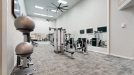 Located on Hickory Club Drive in Nashville, TN, this fitness center offers a generously sized, thoughtfully designed exercise space ideal for a comprehensive workout experience. The facility features soaring ceilings with large skylight panels and oversized ceiling fans that keep the environment bright and well-ventilated throughout. The main strength training area is outfitted with a wide array of cable machines, selectorized weight stations, a dumbbell rack with a full range of free weights, and a flat bench — all arranged to maximize floor space and ease of movement. Full-length mirrors line the walls, enhancing the sense of openness and allowing users to monitor their form. Wall-mounted flat-screen televisions are positioned throughout the space for entertainment and instruction. A mezzanine-level cardio section houses stationary bikes and rowing machines, each paired with individual mounted screens, accessible via a staircase with elegant white railings and a dark wood handrail. Multi-level padded platforms provide versatile space for stretching and functional training. The neutral color palette, comprising white walls and grey patterned carpet, creates a clean, modern aesthetic that makes the entire facility feel welcoming and professional.