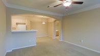 This single-floor residence offers an open-concept layout that promotes a comfortable and connected living experience. The main living area features plush carpeting and is equipped with a ceiling fan, providing both comfort and functionality. Crown molding runs throughout the space, adding a refined architectural detail that enhances the overall aesthetic.

A graceful arched passageway serves as the defining architectural feature, elegantly connecting the living area to the kitchen. The kitchen is outfitted with light wood cabinetry and a raised breakfast bar with a light-toned countertop, providing both additional seating and a natural divide between the kitchen and living spaces. A white refrigerator and a stylish pendant light above the bar round out the kitchen's practical features.

A second arched opening leads to an additional room, likely a bedroom or utility space, with built-in cabinetry visible beyond — suggesting ample storage solutions. The neutral, warm beige tones used throughout the walls and flooring create a cohesive and inviting ambiance. Generous baseboards and recessed HVAC venting in the ceiling further speak to the thoughtful construction of this well-appointed, single-story home.